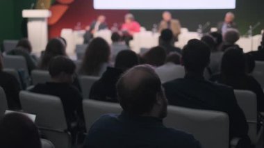 Resmi bir ortamda panel konuşmacılarını dinleyen insanlar, ilgi çekici ve öğrenici.