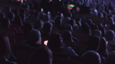 Kalabalık seyirciler, canlı sahne performansını izlerken arkadan çekiliyor, teknoloji kullanımını vurguluyorlar.