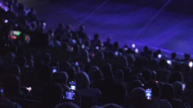 Kalabalık, sahne ışıklarıyla aydınlatılmış akıllı telefonlarla kayıt yaparken bir performans izliyor. Modern teknoloji çağındaki canlı etkinliklerde kalabalığın ilgisini çeker..