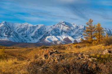 Sonbahar ağaçları ve mavi gökyüzünün altındaki engebeli arazisiyle sersemletici karlı dağ sıraları.
