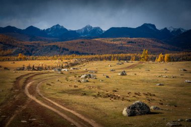 Asfaltsız yolu, dağınık kayaları, ormanlık tepeleri ve bulutlu gökyüzünün altındaki uzak dağları olan engin bir manzara. Sonbahar manzarasının yansıması ve sakin bir çevre..