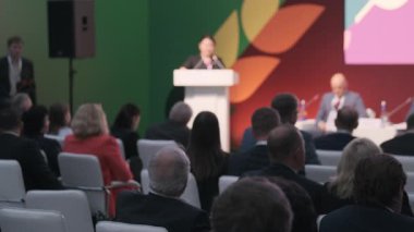 Konuşmacılar, seyirciler, beyaz sandalyeler ve parlak arkaplanlarla bir konferansta bireylerin toplanması.