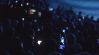 Karanlık bir mekanda akıllı telefonlarıyla sahne performansını kaydeden bir seyircinin siluetleri..