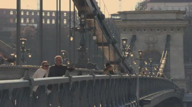 Budapeşte, Macaristan - 2 Nisan 2025: Günbatımının ılık ışığı altında Budapeşte Zincir Köprüsü 'nde yürüyüş yapan çift. Köprü korkulukları ve sütunlarının tarihi mimari ayrıntıları şehir cazibesini yaratır 