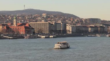 Budapeşte 'nin şehir manzarasında nehirde yüzen bir tekne, önde gelen binalar ve arka planda tepe manzarası var. Güneşli bir günde şehir yaşamını, turizmi ve mimari güzelliği yakalar..