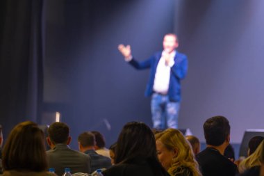 Bir seminerde izleyiciler parlak bir sahnede bir konuşmacıyı dikkatle izliyorlar. Profesyonel iletişim kavramı.
