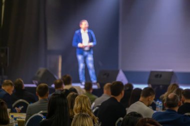 Konuşmacı sahnede duruyor. Bir iş etkinliği sırasında meşgul bir seyirciye sunum yapıyor..