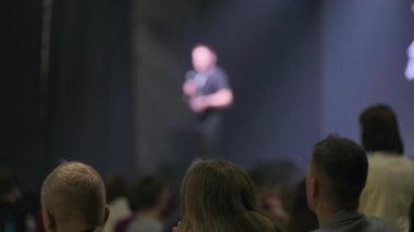 Kalabalık, sahnede odaklanmış ışık altında konuşma yapan bir sunucuyu dinlemek için toplandı..