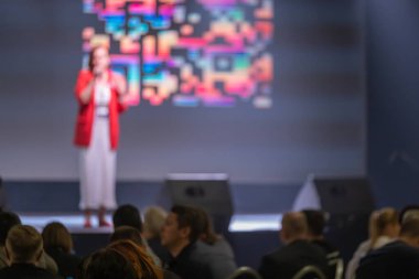 Konferans konuşmacısı sahnede sunum yapıyor. Seyirci izliyor, arkaplanda renkli görüntüler var.