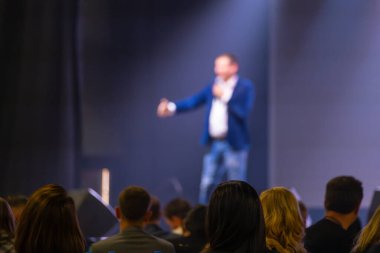 Sunucu sahnede bulanık bir odak ile konuşuyor, bir konferans ortamında izleyicilere hitap ediyor.