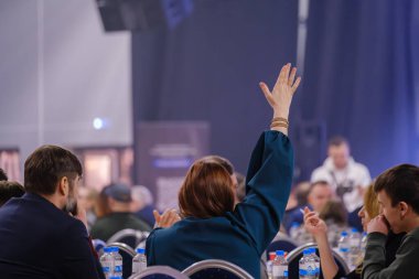 Toplantıya katılan bir grup insan, katılmak için elini kaldıran bir kişi. Sahne aktif tartışma ve eğitim ortamını, takım çalışması ve iletişimi resmediyor.