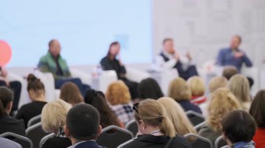 Seyirciler, parlak bir mekandaki profesyonel bir toplantı sırasında çeşitli panelleri dinliyorlar.