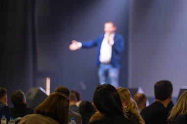 Bir konferans ortamında özenli bir seyirciye sahnede konuşma yapan bir adam.