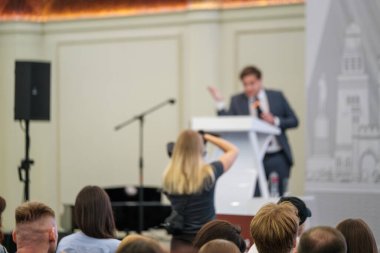 Resmi bir ortamda bir iş konferansı dinleyicisine seslenen konuşmacı, olayı kaydeden bir fotoğrafçıyla.