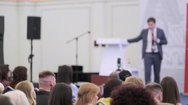 Bir sunucu, resmi bir konferans ortamında izleyiciyle iletişim kuran bir mikrofon tutuyor..