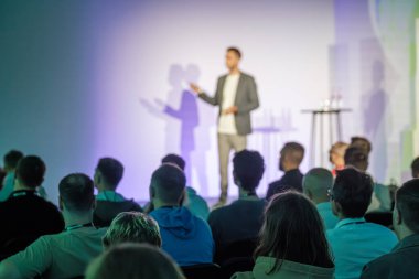 Seyirciler, iyi ışıklandırılmış mekanda seminer sırasında sahnede konuşma yapan sunucuyu dinliyorlar.