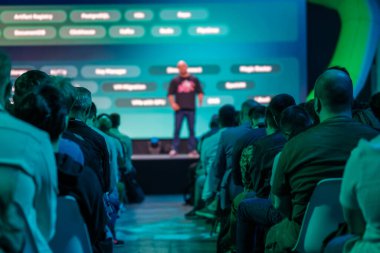 Bir konferans sırasında bir konuşmacı oturmakta olan bir seyirciye hitap eder. Mavi ışıklandırma ve dijital sunum ekranları modern ve çekici bir atmosfer yaratıyor..