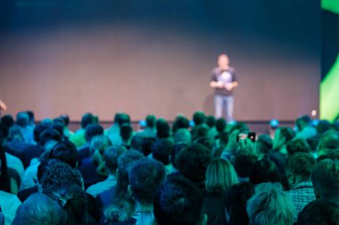 Bir iş konferansına katılanlar parlak bir sahnede bir konuşmacıyı dinliyorlar..