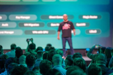 Görünür bir seyirci ve sahnede sunum yapan bir konuşmacıyla konferans sahnesi. Seyircilerin dikkatini ve profesyonel etkinlik atmosferini vurgular.