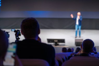 Sahnedeki konuşmacı, kısmen görülen seyircilere görüntüleme anları için yerleştirilen kayıt ekipmanlarını sunar