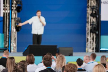 Sunucu, modern ışıklandırma ve kurulum ile mavi sahnede özenli seyircilere sesleniyor