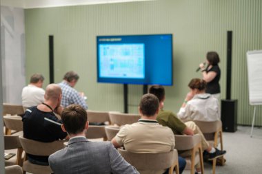 Konferans odasında oturan katılımcılar bir ekran sunumu kullanarak sunum yapan kadını dinliyorlar.