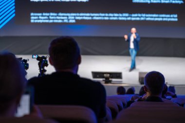 Konferans salonundaki izleyiciler sahnedeki bir hoparlöre odaklanırken, kameralar kayıt yapıyor..