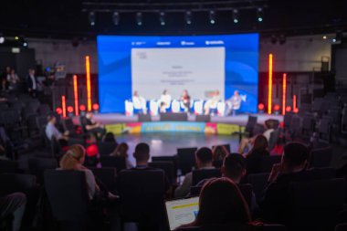Oturan izleyiciler, büyük ekran ve sahne paneli ile konferansın bulanık görünümü.