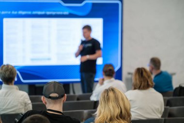 Bir konferansta iş ya da eğitim sunumunu sergileyen bir konuşmacıyı dinleyen seyirciler.