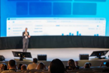 Hoparlör sahnede seyirciye sunar, büyük ekranda grafikler görünür.