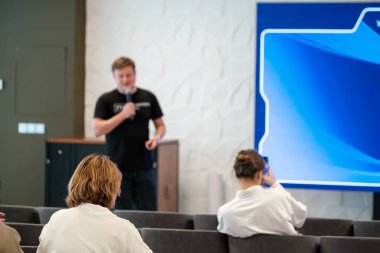 İzleyiciler mikrofon ve ekran kullanarak sunum yapan bir hoparlörü dinliyorlar.