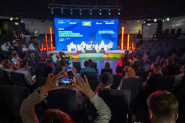 İş etkinliği, sunum sırasında izleyicilerin akıllı telefonlarla sahnede panel tartışmasını kaydettiğini gösteriyor. Modern bir mekanda etkileşimli iş ve ağ kurma vesilesi.