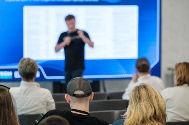 Büyük ekranlı bir konferans sunucusunu dinleyen odaklanmış seyirciler.