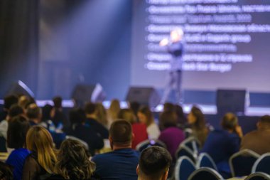 Konferansta, loş ışıklandırmalı oditoryumdaki seyircilere sunum yapan konuşmacı.