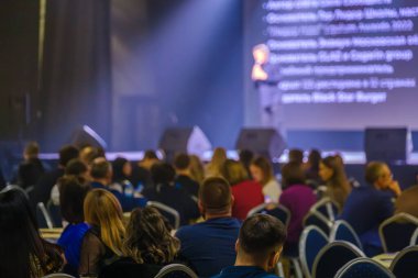 Kalabalık konferansta motivasyon konuşmasına katılıyor. Hoparlör sahnede seyirciye yumuşak ışık altında sesleniyor.