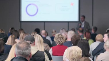 Kalabalık bir konferans salonu, önde bir konuşmacı dinliyor, projektör ekranı grafikleri gösteriyor, profesyonel öğrenme, bağlanma ve kurumsal bir etkinlikte işbirliği kaydediyor.