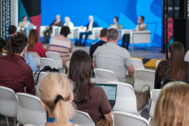 Modern konferansta çeşitli konuşmacıların parlak mavi sahne arkaplanı, odak noktası ve profesyonel enerjiyi yakalayan seyircilere seslendiği bir panel tartışması