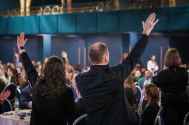 Konferanstaki profesyoneller, kalabalık etkinlik mekanında katılımcılar arasında yer alan katılım ve enerjiyi gösteren sunum sırasında coşkuyla el kaldırıyorlar.