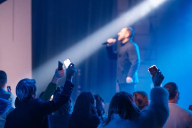 Canlı konserdeki kalabalık, şarkıcının mavi spot ışıkları altında, hayranlarının telefonları çekmesiyle alkış tutuyor.