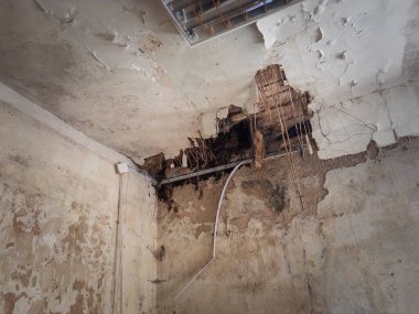 Çatlak alçılı hasarlı oda tavandaki büyük delikler paslanmış borular çürüme hissi yaratıyor ve tamamlanmamış binada ihmal var.