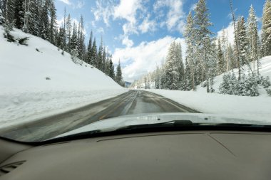 Arkasında mavi gökyüzü olan bir ormanda, karla kaplı dağların içinden geçen bir arabanın görüntüsü.