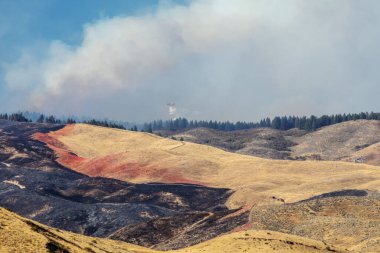 Görünür ateş hatları, duman ve Boise Idaho yakınlarındaki 2024 Vadisi Yangınından kalma yanmış manzaralı dağlık bir bölgede yangın söndürme çabalarını gösteren dramatik bir hava sahnesi.