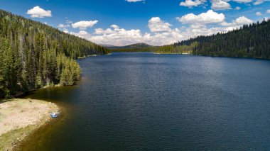 Sakin suları ve gür çam ormanları olan sakin bir dağ gölü canlı mavi bir gökyüzü altında tüylü bulutlarla huzurlu bir doğal geri çekilme sunuyor..
