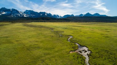 Stanley Idaho 'daki dolambaçlı bir deresi ve karlı dağları olan yemyeşil bir arazinin geniş bir hava görüntüsü. Doğa ve seyahat temaları için mükemmel.
