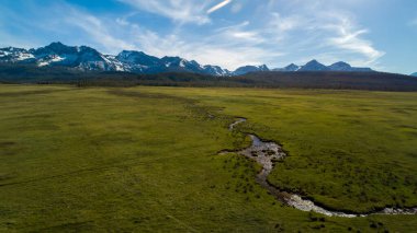 İçinden nehir akan geniş, açık bir alan. Gökyüzü açık ve mavidir ve arka plandaki dağlar karla kaplıdır. Huzurlu ve huzurlu bir sahne.