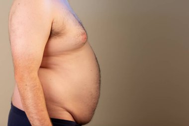 Kocaman göbekli bir adam bir duvarın önünde duruyor. Adamın karnı dışarı çıkıyor ve mavi pantolon giyiyor.