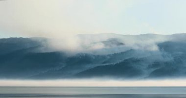 Önünde bir su kütlesi olan sisli bir dağ sırası uzaktan görülebilir. Huzurlu ve huzurlu bir manzara, sisli atmosfer sükunet duygusunu artırıyor.