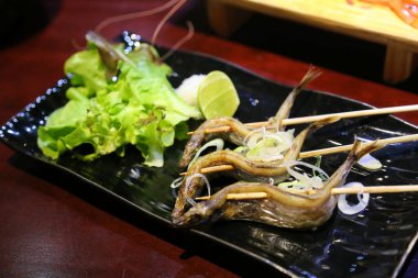 Japon mutfağının sığ odak noktası, şiş üstünde ızgara Shishamo balığı. Dikdörtgen siyah bir tabakta sebzeler ve limon dilimleriyle güzelce düzenlenmiş. Bir Japon restoranında..