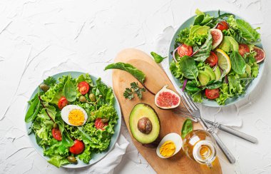 Fresh green lettuce salads with mixed vegetables, egg and fig on white table background clipart