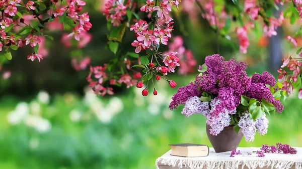 bouquet of lilacs in a vase on the table. Against the backdrop of cherry blossoms with beautiful bokeh, background and wallpaper for design with empty space to insert text